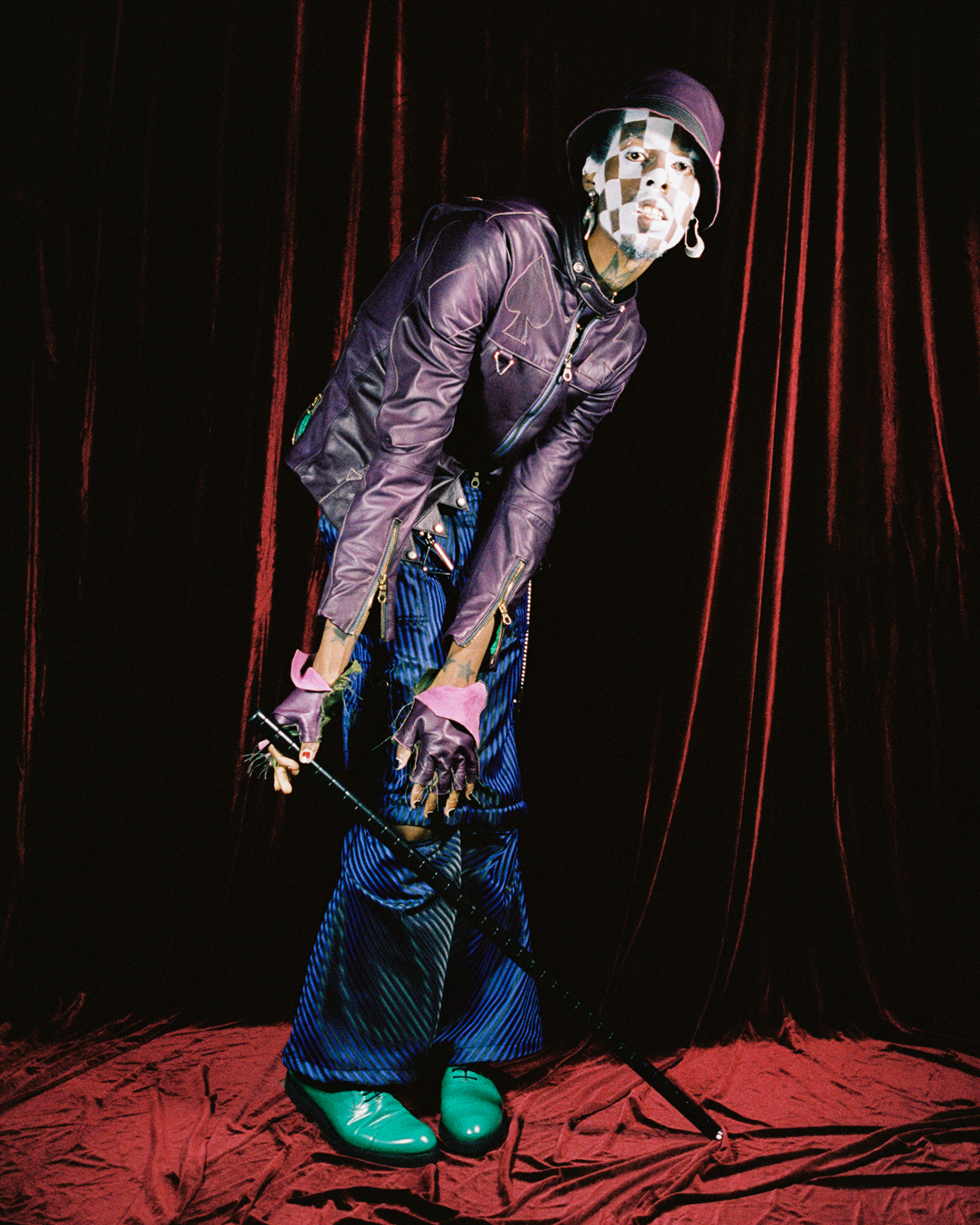 Deep-skined model with white checkered makeup crouched with a cane wearing a pruple jacket and blue pants against a red velvet background